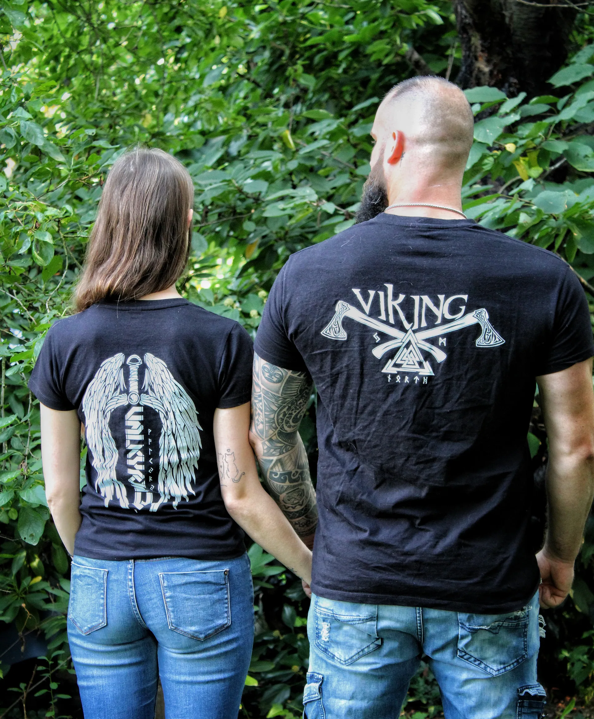 Paar von hinten Hand in Hand im Wald, Rückenmotive: Valkyria-Flügel bei der Frau, Wikinger-Äxte beim Mann
