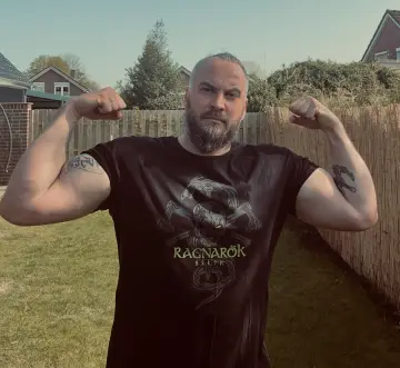 Tätowierter Mann zeigt Bizepse in einem North Legendary Ragnarök-Shirt mit Drachen-Motiv im Garten